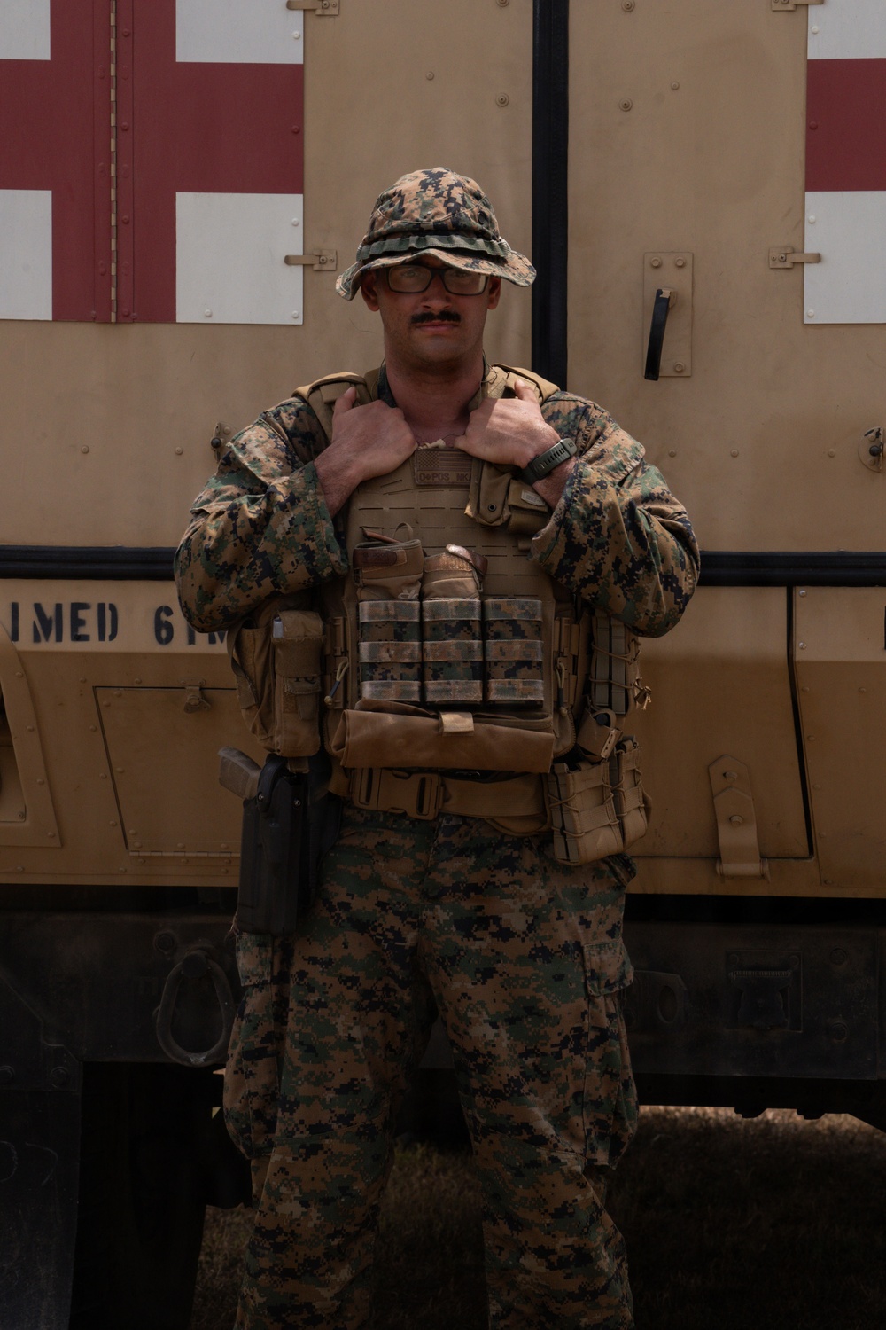 A male service member in U.S. Marine Corps MARPAT woodland/desert digital camouflage stands in front of a military vehicle marked with a red cross (medical vehicle). He is wearing full combat gear including a plate carrier vest with magazine pouches, a boonie hat, glasses, and a mustache. The setting appears to be an outdoor military environment, likely a forward operating base or field exercise.