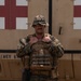 A male service member in U.S. Marine Corps MARPAT woodland/desert digital camouflage stands in front of a military vehicle marked with a red cross (medical vehicle). He is wearing full combat gear including a plate carrier vest with magazine pouches, a boonie hat, glasses, and a mustache. The setting appears to be an outdoor military environment, likely a forward operating base or field exercise.