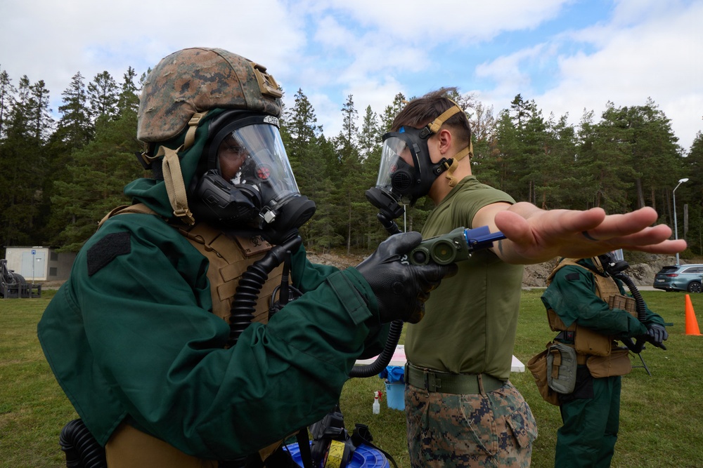 Military personnel in CBRN (Chemical, Biological, Radiological, and Nuclear) protective gear are conducting a decontamination or detection exercise outdoors. One soldier in full green protective suit and combat helmet is using a handheld detection device to scan another soldier's outstretched arm. The setting appears to be a training exercise at a military installation surrounded by pine trees.