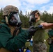 Military personnel in CBRN (Chemical, Biological, Radiological, and Nuclear) protective gear are conducting a decontamination or detection exercise outdoors. One soldier in full green protective suit and combat helmet is using a handheld detection device to scan another soldier's outstretched arm. The setting appears to be a training exercise at a military installation surrounded by pine trees.