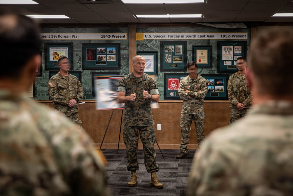 A military briefing or presentation is taking place in what appears to be a Special Forces heritage room or museum. A central figure in Marine Corps MARPAT camouflage uniform is addressing a group of soldiers, with framed historical displays visible on the wall behind him. The wall displays reference U.S. Special Forces history including Korean War and Southeast Asia operations from 1942 to present.