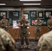 A military briefing or presentation is taking place in what appears to be a Special Forces heritage room or museum. A central figure in Marine Corps MARPAT camouflage uniform is addressing a group of soldiers, with framed historical displays visible on the wall behind him. The wall displays reference U.S. Special Forces history including Korean War and Southeast Asia operations from 1942 to present.