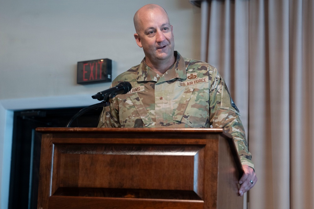A U.S. Air Force officer is speaking at a wooden podium with a microphone. The individual wears an OCP (Operational Camouflage Pattern) uniform with a name tape partially reading 'LIAMS' (likely 'WILLIAMS'), a single star insignia indicating the rank of Brigadier General, and a 'U.S. AIR FORCE' tape. The setting appears to be an indoor event or ceremony venue.