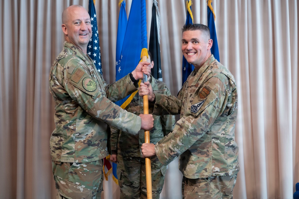 Two U.S. Air Force personnel in OCP (Operational Camouflage Pattern) uniforms are participating in a formal guidon/flag passing ceremony, likely a change of command. Both individuals are smiling while jointly holding a wooden staff with a yellow tassel, indicating a transfer of unit colors. A third service member stands in the background near multiple flags including the U.S. flag and Air Force flag.