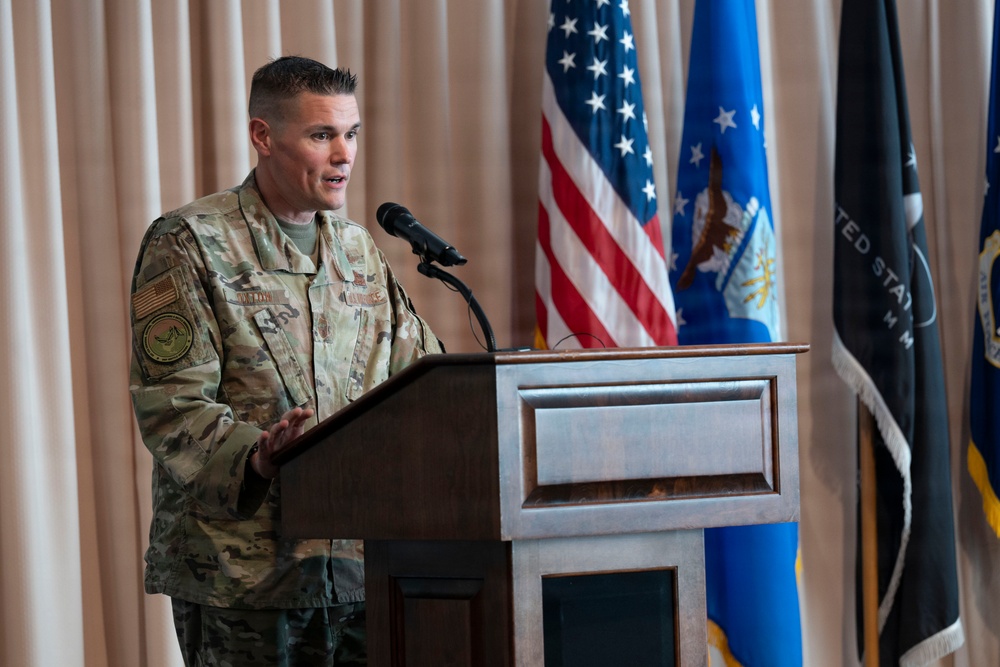 A military officer in U.S. Air Force OCP (Operational Camouflage Pattern) uniform is speaking at a wooden podium with a microphone. The setting appears to be a formal military ceremony or briefing, with multiple flags visible in the background including the U.S. flag and what appears to be Air Force and command flags. The backdrop consists of beige/cream curtains in an indoor venue.