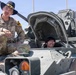 A U.S. Army soldier wearing a cavalry Stetson hat and OCP camouflage uniform sits atop a military armored vehicle, speaking with a smiling civilian who is seated inside the vehicle's open hatch. The soldier's name tape reads 'WHITE' and he wears a 1st Cavalry Division patch on his left shoulder. This appears to be a public outreach or demonstration event where civilians are given access to military equipment.