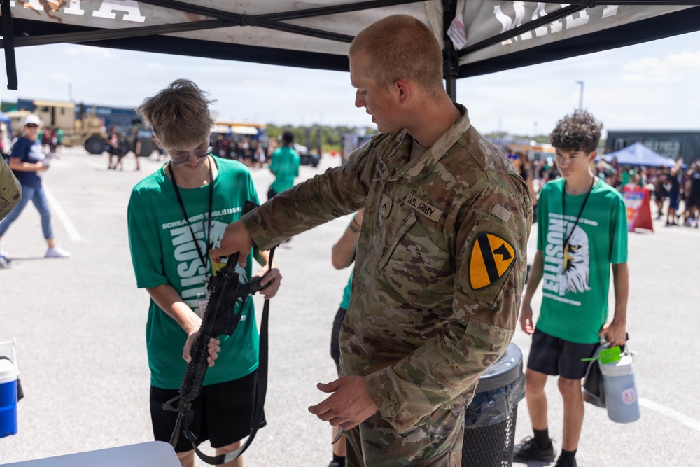 A U.S. Army soldier wearing 1st Cavalry Division insignia is demonstrating a rifle to a young person at what appears to be a military recruitment or public outreach event. Several other young people in matching green 'Screaming Eagles' band shirts are present under a canopy tent. The setting appears to be an outdoor military expo or air show with military vehicles visible in the background.
