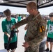 A U.S. Army soldier wearing 1st Cavalry Division insignia is demonstrating a rifle to a young person at what appears to be a military recruitment or public outreach event. Several other young people in matching green 'Screaming Eagles' band shirts are present under a canopy tent. The setting appears to be an outdoor military expo or air show with military vehicles visible in the background.