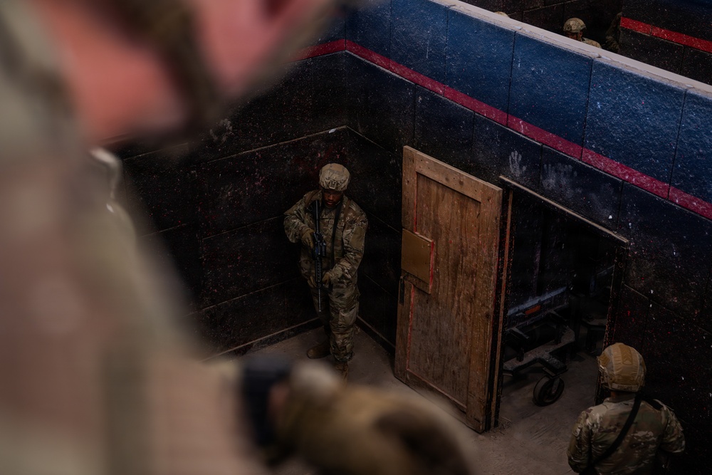 The image shows military personnel in combat gear conducting what appears to be a close-quarters battle (CQB) or room-clearing training exercise. The scene is photographed from an elevated angle, showing soldiers in multicam/OCP uniforms with helmets and rifles positioned near a wooden door in a dark-walled training facility. The environment resembles a military shoot house or urban operations training center.
