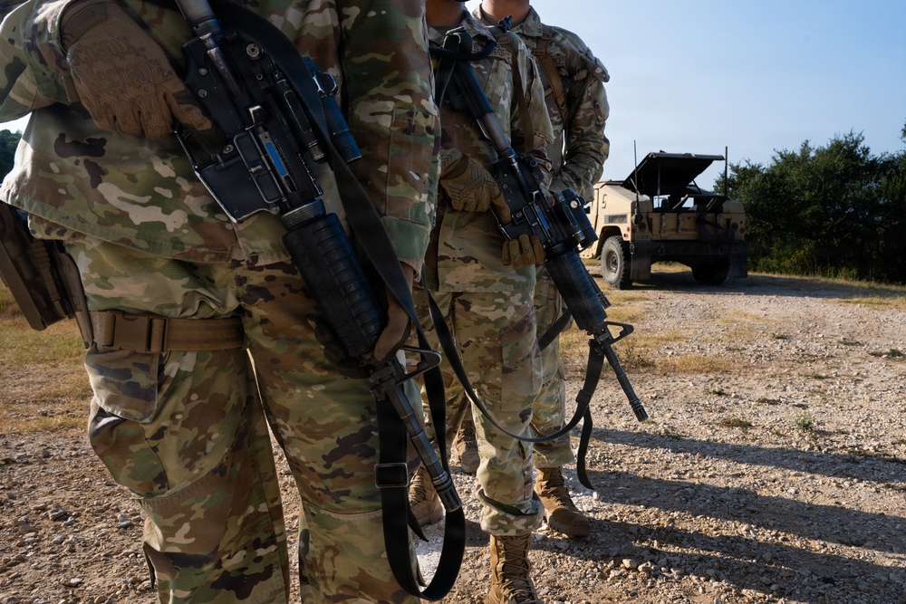 Two military personnel in MultiCam/OCP camouflage uniforms are standing on a gravel surface while carrying rifles slung across their bodies. A military tactical vehicle is visible in the background on what appears to be a training area or field environment. The image is cropped at torso level, obscuring the subjects' faces.