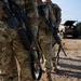 Two military personnel in MultiCam/OCP camouflage uniforms are standing on a gravel surface while carrying rifles slung across their bodies. A military tactical vehicle is visible in the background on what appears to be a training area or field environment. The image is cropped at torso level, obscuring the subjects' faces.