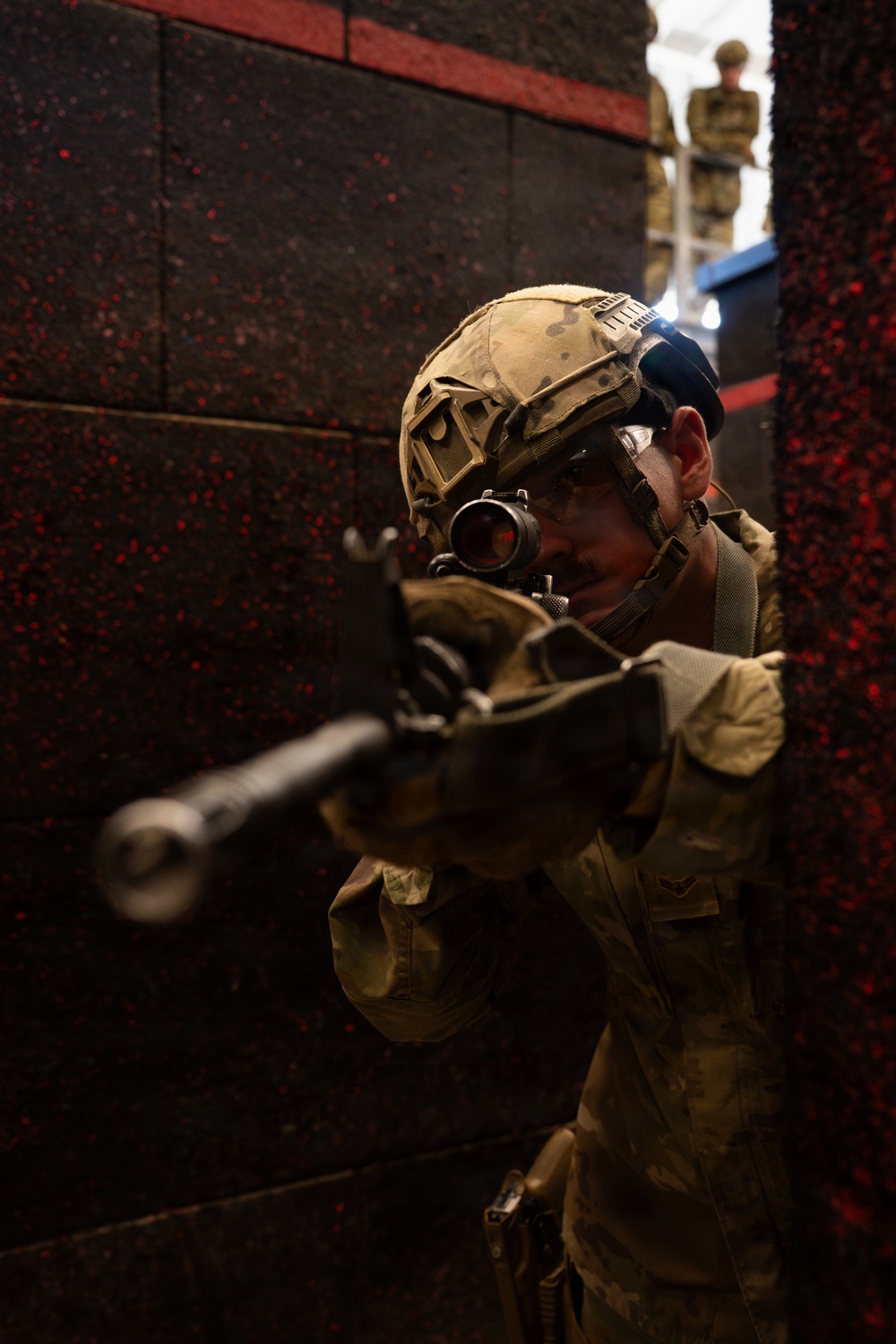 A soldier in full tactical gear is positioned against a dark wall, aiming a rifle with an optical scope directly toward the camera. The foreground figure is in a combat-ready stance, using the wall for cover. A second uniformed individual is visible in the background through a gap between structures.