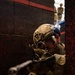 A soldier in full tactical gear is positioned against a dark wall, aiming a rifle with an optical scope directly toward the camera. The foreground figure is in a combat-ready stance, using the wall for cover. A second uniformed individual is visible in the background through a gap between structures.