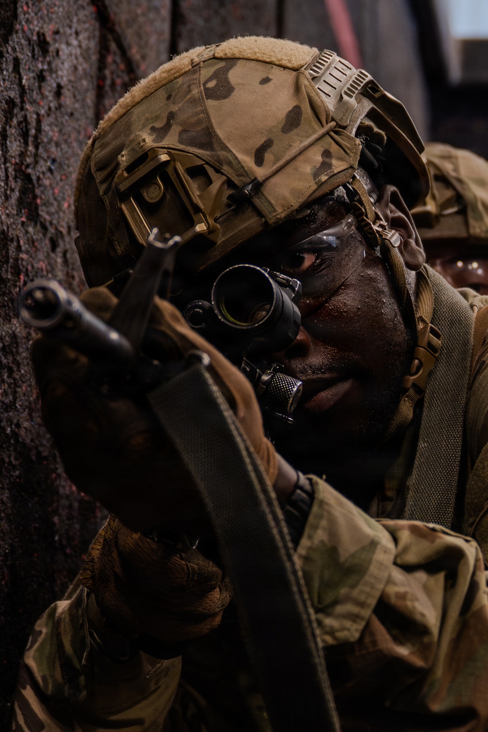 A soldier in full tactical gear is seen aiming down the optic sight of a rifle while positioned against a dark wall or structure. The individual is wearing a multicam-patterned helmet with a night-vision device mount and tactical gloves. The image captures a close-up, high-intensity moment consistent with a military training exercise or operational scenario.