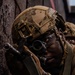 A soldier in full tactical gear is seen aiming down the optic sight of a rifle while positioned against a dark wall or structure. The individual is wearing a multicam-patterned helmet with a night-vision device mount and tactical gloves. The image captures a close-up, high-intensity moment consistent with a military training exercise or operational scenario.