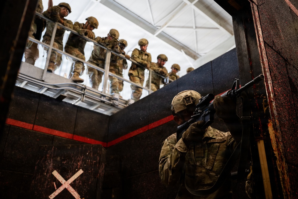 A U.S. Air Force service member in full combat gear is positioned in a doorway aiming a rifle during what appears to be a military training exercise. A group of soldiers in camouflage uniforms and helmets observe from an elevated platform above. The setting appears to be a purpose-built military training facility with marked walls and industrial infrastructure.