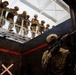 A U.S. Air Force service member in full combat gear is positioned in a doorway aiming a rifle during what appears to be a military training exercise. A group of soldiers in camouflage uniforms and helmets observe from an elevated platform above. The setting appears to be a purpose-built military training facility with marked walls and industrial infrastructure.