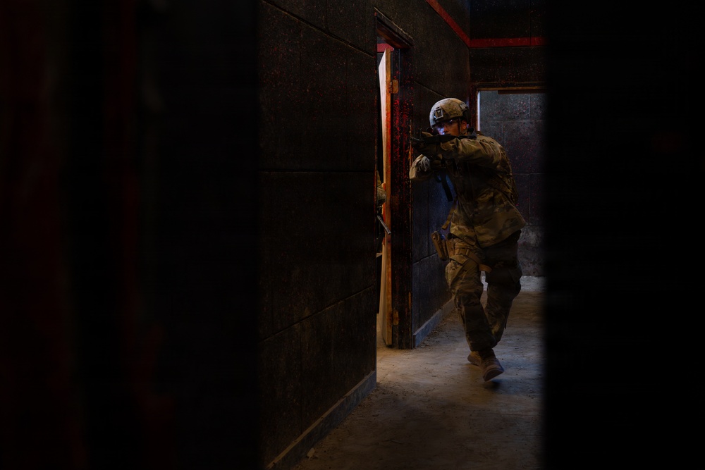 A soldier in full combat gear is conducting a room-clearing or building-entry operation in a dark, confined corridor. The individual is in a tactical stance, aiming a weapon while moving through a doorway. The environment appears to be a training facility or urban operations structure based on the controlled lighting and constructed walls.
