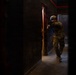 A soldier in full combat gear is conducting a room-clearing or building-entry operation in a dark, confined corridor. The individual is in a tactical stance, aiming a weapon while moving through a doorway. The environment appears to be a training facility or urban operations structure based on the controlled lighting and constructed walls.