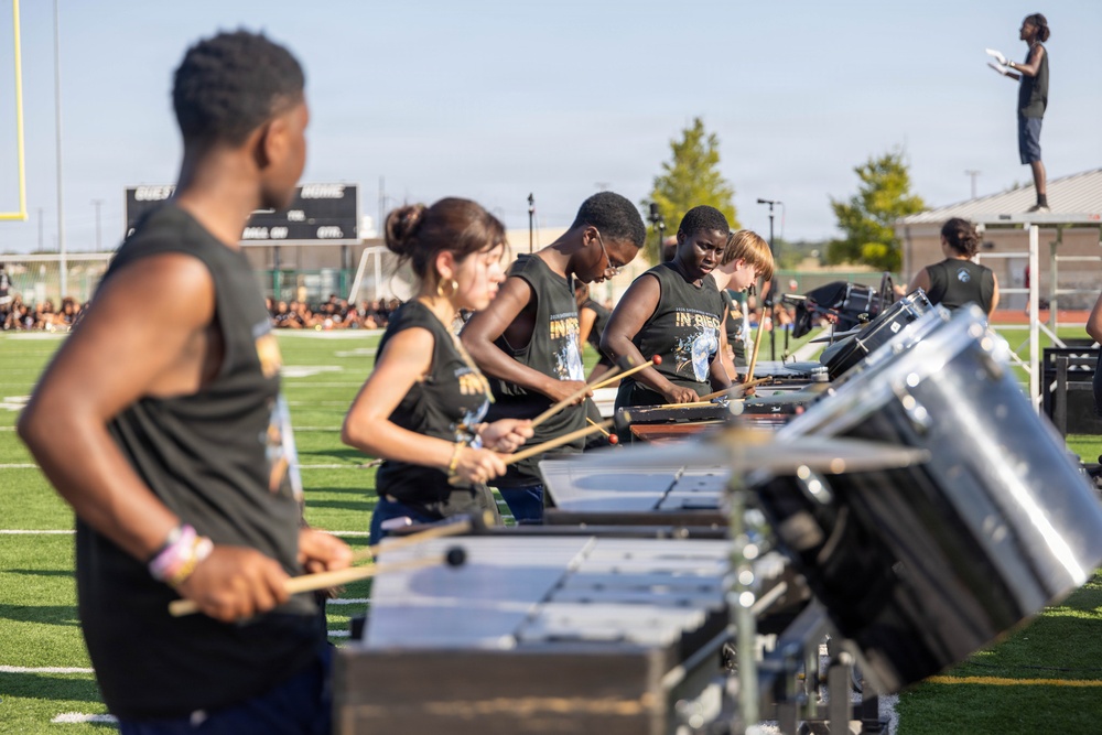 A percussion ensemble performs on a football field, with multiple musicians playing marimbas and snare drums in a line formation. The performers are wearing matching dark sleeveless shirts, suggesting an organized marching band or drum corps event. A conductor or director is visible standing elevated on a platform in the background, overseeing the performance.