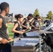 A percussion ensemble performs on a football field, with multiple musicians playing marimbas and snare drums in a line formation. The performers are wearing matching dark sleeveless shirts, suggesting an organized marching band or drum corps event. A conductor or director is visible standing elevated on a platform in the background, overseeing the performance.