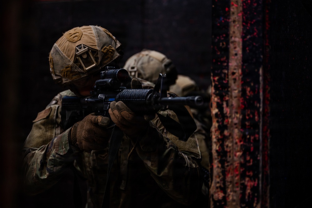 Two military personnel in full tactical gear are conducting what appears to be a close-quarters battle (CQB) training exercise or operation in a dark, confined environment. The foreground soldier is aiming an M4-style carbine rifle equipped with an optic sight through a doorway or opening. Both soldiers wear multicam camouflage uniforms and tactical helmets with mounted accessories.