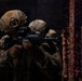 Two military personnel in full tactical gear are conducting what appears to be a close-quarters battle (CQB) training exercise or operation in a dark, confined environment. The foreground soldier is aiming an M4-style carbine rifle equipped with an optic sight through a doorway or opening. Both soldiers wear multicam camouflage uniforms and tactical helmets with mounted accessories.