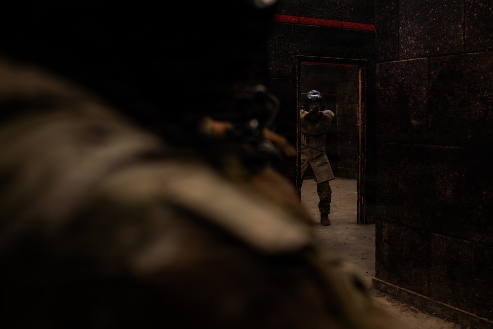 The image depicts two armed individuals in tactical gear inside a dark, industrial-looking structure. One person is in the foreground (blurred/out of focus) appearing to aim a weapon, while a second person is visible in the background standing in a doorway also in a shooting stance. The setting and equipment suggest a military or law enforcement training exercise.