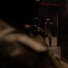 The image depicts two armed individuals in tactical gear inside a dark, industrial-looking structure. One person is in the foreground (blurred/out of focus) appearing to aim a weapon, while a second person is visible in the background standing in a doorway also in a shooting stance. The setting and equipment suggest a military or law enforcement training exercise.