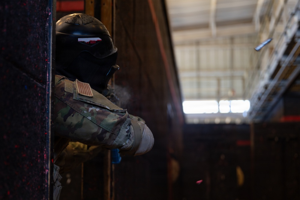 A military personnel wearing full protective gear including a black helmet with face shield and camouflage uniform is positioned at the edge of a doorway or wall, peering around a corner. The individual has a US flag patch visible on their left shoulder, indicating US military affiliation. The setting appears to be an indoor training facility or hangar environment.