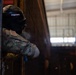 A military personnel wearing full protective gear including a black helmet with face shield and camouflage uniform is positioned at the edge of a doorway or wall, peering around a corner. The individual has a US flag patch visible on their left shoulder, indicating US military affiliation. The setting appears to be an indoor training facility or hangar environment.