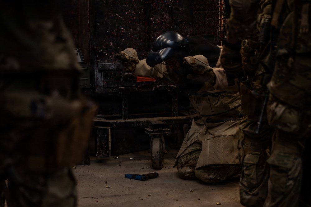 A military or law enforcement personnel wearing a bomb disposal or protective helmet and camouflage uniform is kneeling and working on or inspecting a dark metal object or vehicle component. The scene appears to be an indoor training or operational environment with other uniformed personnel visible in the foreground and background. The surface shows scattered debris and what appears to be paint or residue marks on the dark metal surface.