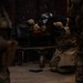 A military or law enforcement personnel wearing a bomb disposal or protective helmet and camouflage uniform is kneeling and working on or inspecting a dark metal object or vehicle component. The scene appears to be an indoor training or operational environment with other uniformed personnel visible in the foreground and background. The surface shows scattered debris and what appears to be paint or residue marks on the dark metal surface.