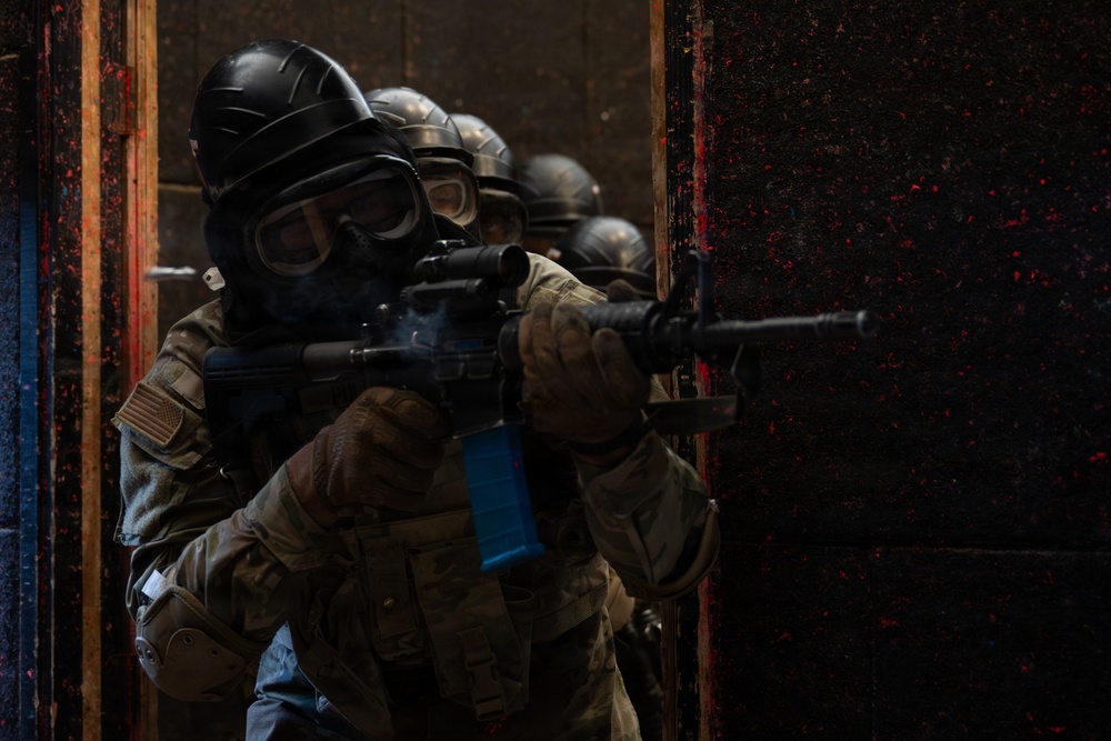 The image shows a tactical team in full protective gear conducting a room-clearing or breach-and-entry drill. Multiple personnel are stacked in formation at a doorway, each armed with rifles and wearing gas masks and helmets. The blue magazine on the lead weapon and paint splatter on the door suggest this is a training exercise using simunition or paintball-type training ammunition.