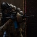 The image shows a tactical team in full protective gear conducting a room-clearing or breach-and-entry drill. Multiple personnel are stacked in formation at a doorway, each armed with rifles and wearing gas masks and helmets. The blue magazine on the lead weapon and paint splatter on the door suggest this is a training exercise using simunition or paintball-type training ammunition.