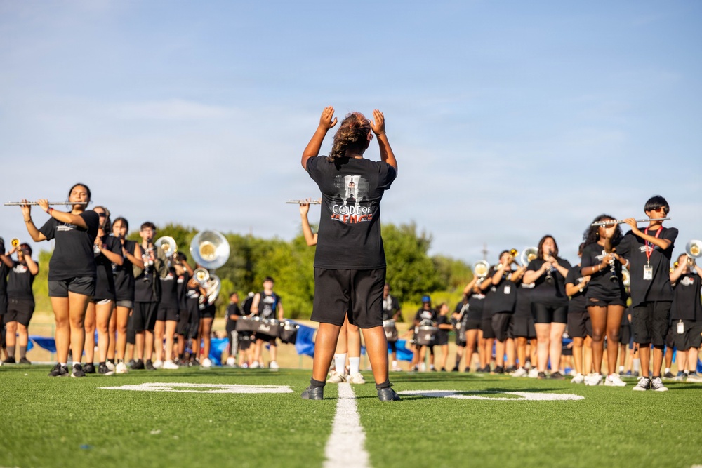 A marching band is rehearsing on a green turf football field under a clear blue sky. A conductor or drum major stands in the foreground with both hands raised, directing the ensemble. Band members dressed in black t-shirts and shorts are spread across the field playing various instruments including flutes, trombones, and sousaphones.