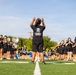 A marching band is rehearsing on a green turf football field under a clear blue sky. A conductor or drum major stands in the foreground with both hands raised, directing the ensemble. Band members dressed in black t-shirts and shorts are spread across the field playing various instruments including flutes, trombones, and sousaphones.