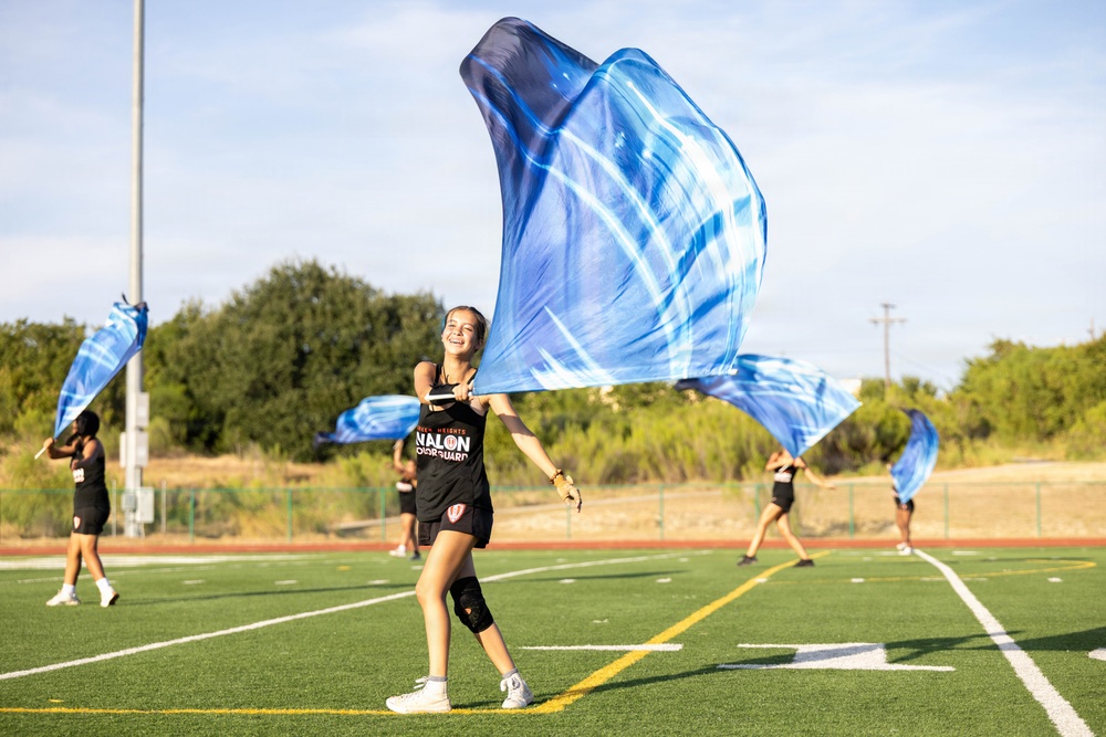 A color guard team is practicing on an outdoor artificial turf football field. The central performer, a smiling young woman wearing a black tank top with 'NALON COLOR GUARD' text, is spinning a large blue and white silk flag. At least two additional performers are visible in the background doing the same activity.