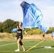 A color guard team is practicing on an outdoor artificial turf football field. The central performer, a smiling young woman wearing a black tank top with 'NALON COLOR GUARD' text, is spinning a large blue and white silk flag. At least two additional performers are visible in the background doing the same activity.