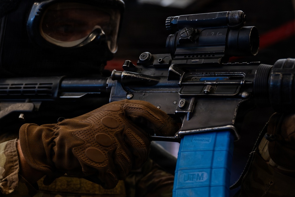 A close-up photograph of a person in tactical gear holding an AR-style rifle with a blue UTM (Ultimate Training Munitions) magazine inserted, indicating a training exercise rather than live-fire combat. The individual is wearing brown tactical gloves, a protective face mask/goggles, and military-style clothing. The weapon is equipped with a magnified optic scope mounted on a rail system.
