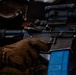 A close-up photograph of a person in tactical gear holding an AR-style rifle with a blue UTM (Ultimate Training Munitions) magazine inserted, indicating a training exercise rather than live-fire combat. The individual is wearing brown tactical gloves, a protective face mask/goggles, and military-style clothing. The weapon is equipped with a magnified optic scope mounted on a rail system.
