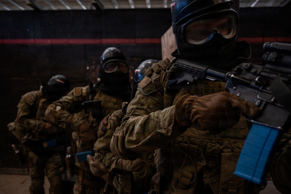 The image shows a group of tactical personnel in full combat gear, including camouflage uniforms, protective goggles, balaclavas, and tactical vests. They are holding rifles with blue-marked magazines, which are commonly used in training exercises to indicate simulated/blank ammunition. The setting appears to be an indoor training facility or range.