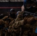 The image shows a group of tactical personnel in full combat gear, including camouflage uniforms, protective goggles, balaclavas, and tactical vests. They are holding rifles with blue-marked magazines, which are commonly used in training exercises to indicate simulated/blank ammunition. The setting appears to be an indoor training facility or range.