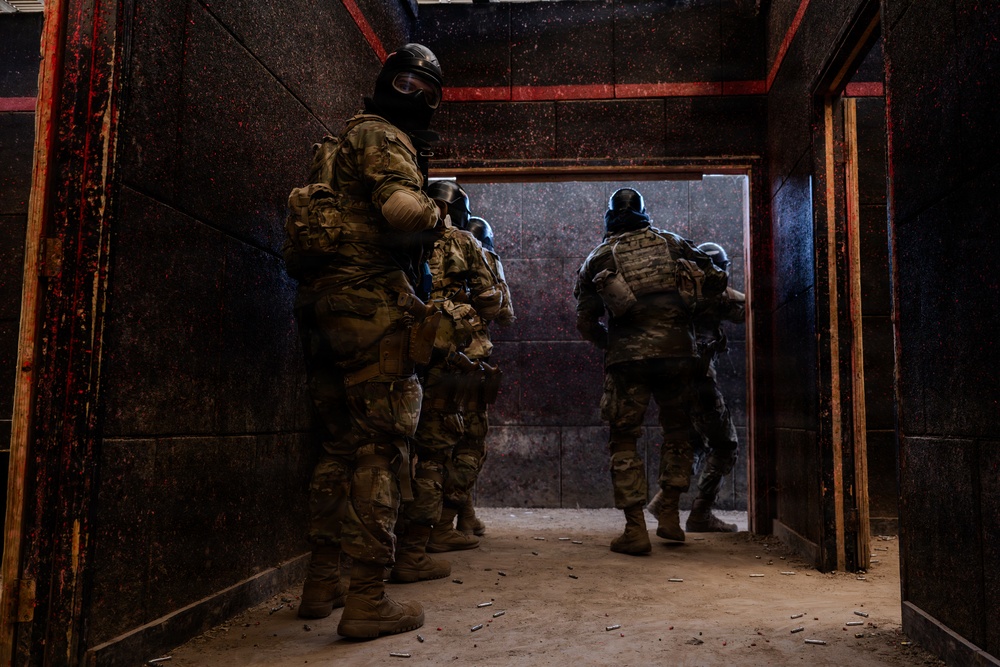 The image shows a group of tactical operators in full military/SWAT gear moving through a dark corridor toward a doorway. The personnel are wearing camouflage uniforms, tactical vests, and black balaclavas/helmets. The setting appears to be a training facility or urban operations environment, with spent shell casings visible on the floor.