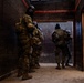 The image shows a group of tactical operators in full military/SWAT gear moving through a dark corridor toward a doorway. The personnel are wearing camouflage uniforms, tactical vests, and black balaclavas/helmets. The setting appears to be a training facility or urban operations environment, with spent shell casings visible on the floor.