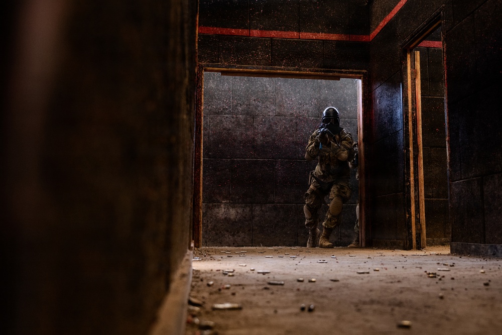 A heavily equipped tactical operator in full combat gear, including a gas mask and body armor, is positioned in a doorway of a dark, deteriorated building. The figure is holding a weapon in a ready position, appearing to conduct a room-clearing or building-entry maneuver. The environment shows signs of significant decay with debris scattered across the floor.