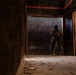 A heavily equipped tactical operator in full combat gear, including a gas mask and body armor, is positioned in a doorway of a dark, deteriorated building. The figure is holding a weapon in a ready position, appearing to conduct a room-clearing or building-entry maneuver. The environment shows signs of significant decay with debris scattered across the floor.