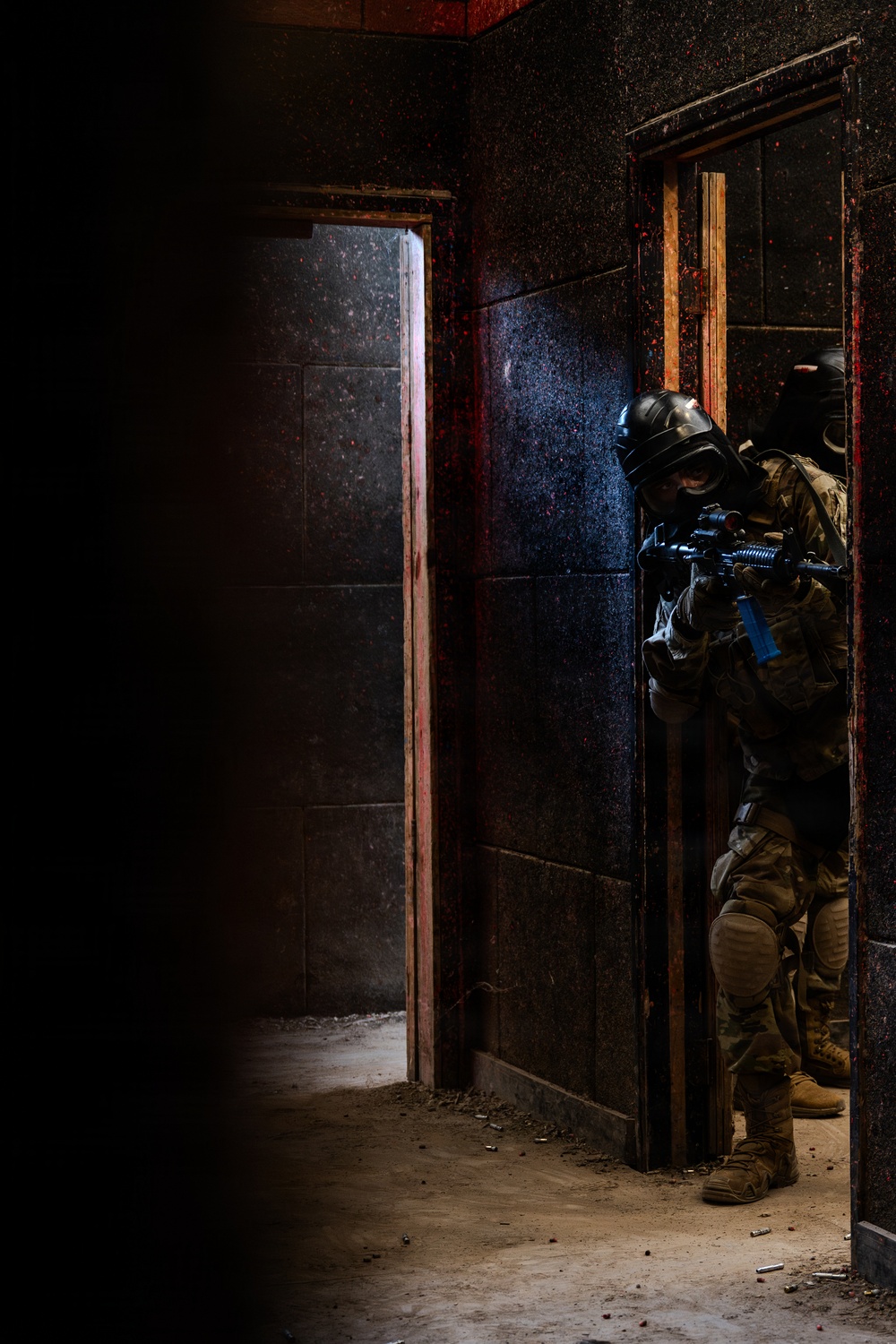 The image depicts two heavily equipped tactical operators in full protective gear conducting a room-clearing or building-entry drill in what appears to be a training facility. The lead operator is positioned at a doorway in a low crouch, aiming a rifle with a blue training barrel indicator. The environment shows a darkened, weathered interior with red paint splatter markings typical of military or law enforcement training structures.
