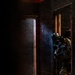 The image depicts two heavily equipped tactical operators in full protective gear conducting a room-clearing or building-entry drill in what appears to be a training facility. The lead operator is positioned at a doorway in a low crouch, aiming a rifle with a blue training barrel indicator. The environment shows a darkened, weathered interior with red paint splatter markings typical of military or law enforcement training structures.