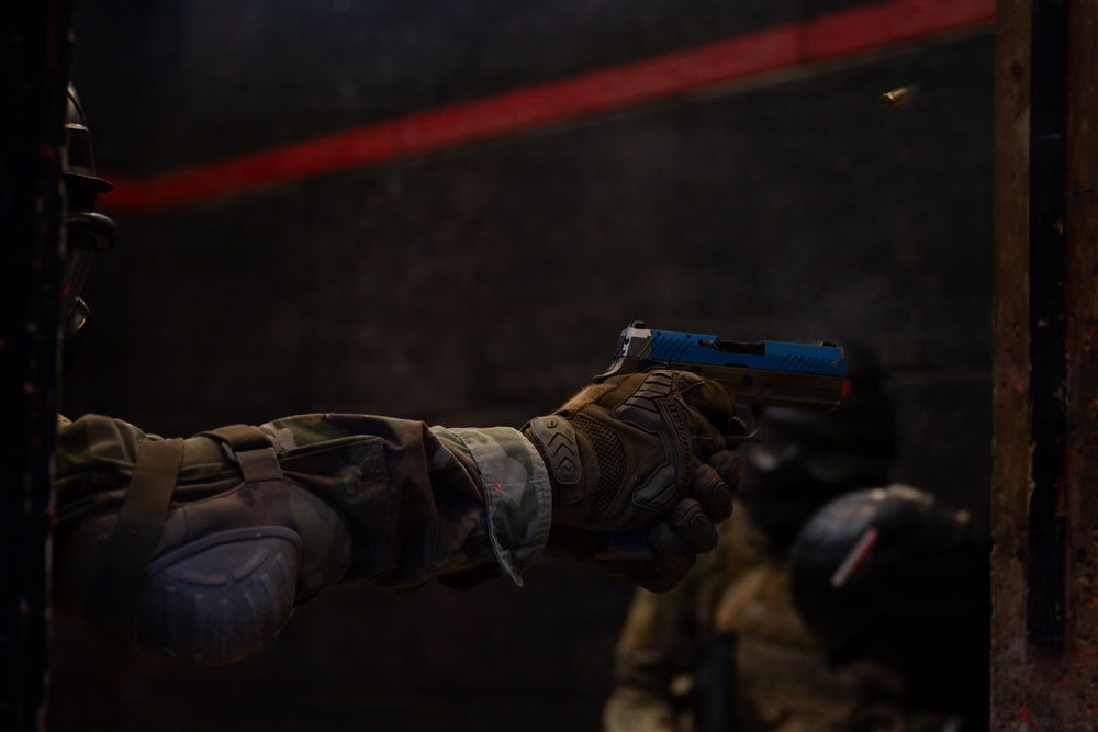 A person in tactical/military gear and camouflage uniform is firing a pistol with a distinctive blue slide in a dark indoor environment. A spent brass casing is visible in the air, indicating the weapon has just been discharged. A second person in tactical gear and helmet is visible in the background.